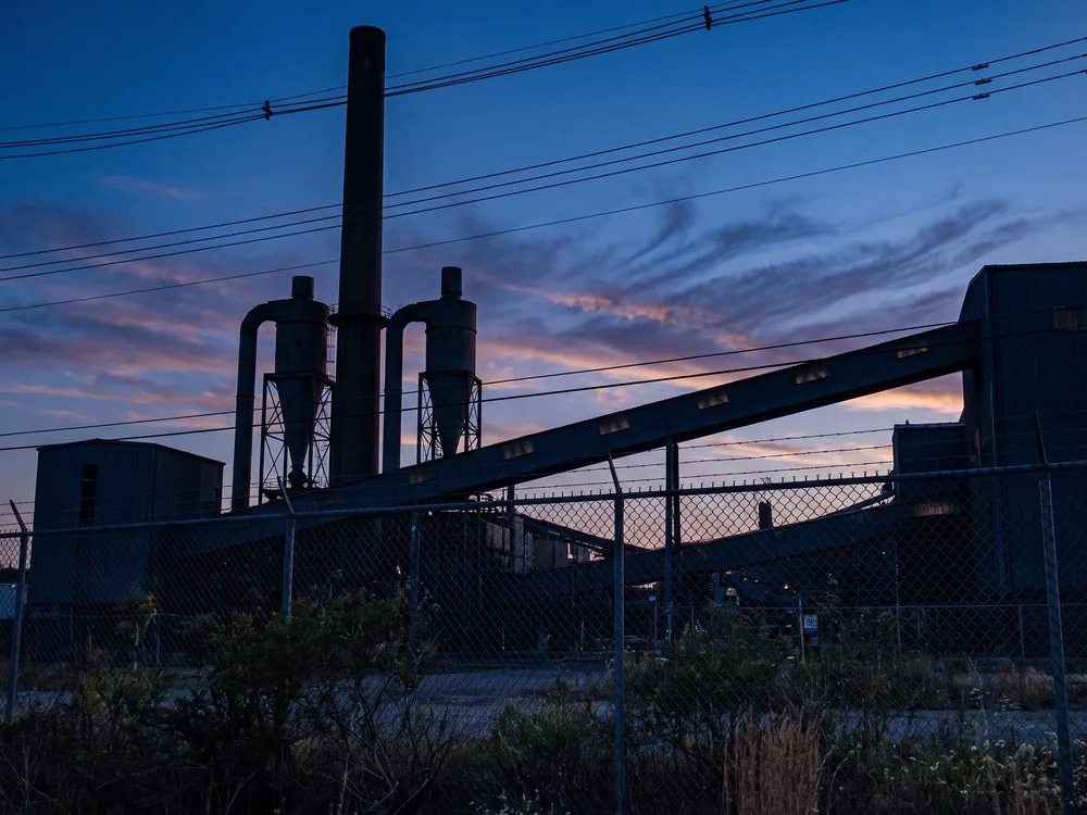 An industrial works at dusk in silhouette: a tall stack, two cyclone separators and a covered conveyor gallery running on a diagonal.